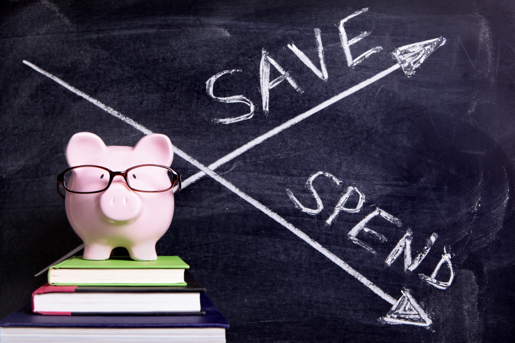 Pink piggy bank with glasses standing on books next to a blackboard with simple spend and save message. Sharp focus on the piggy bank with blackboard slightly blurred.